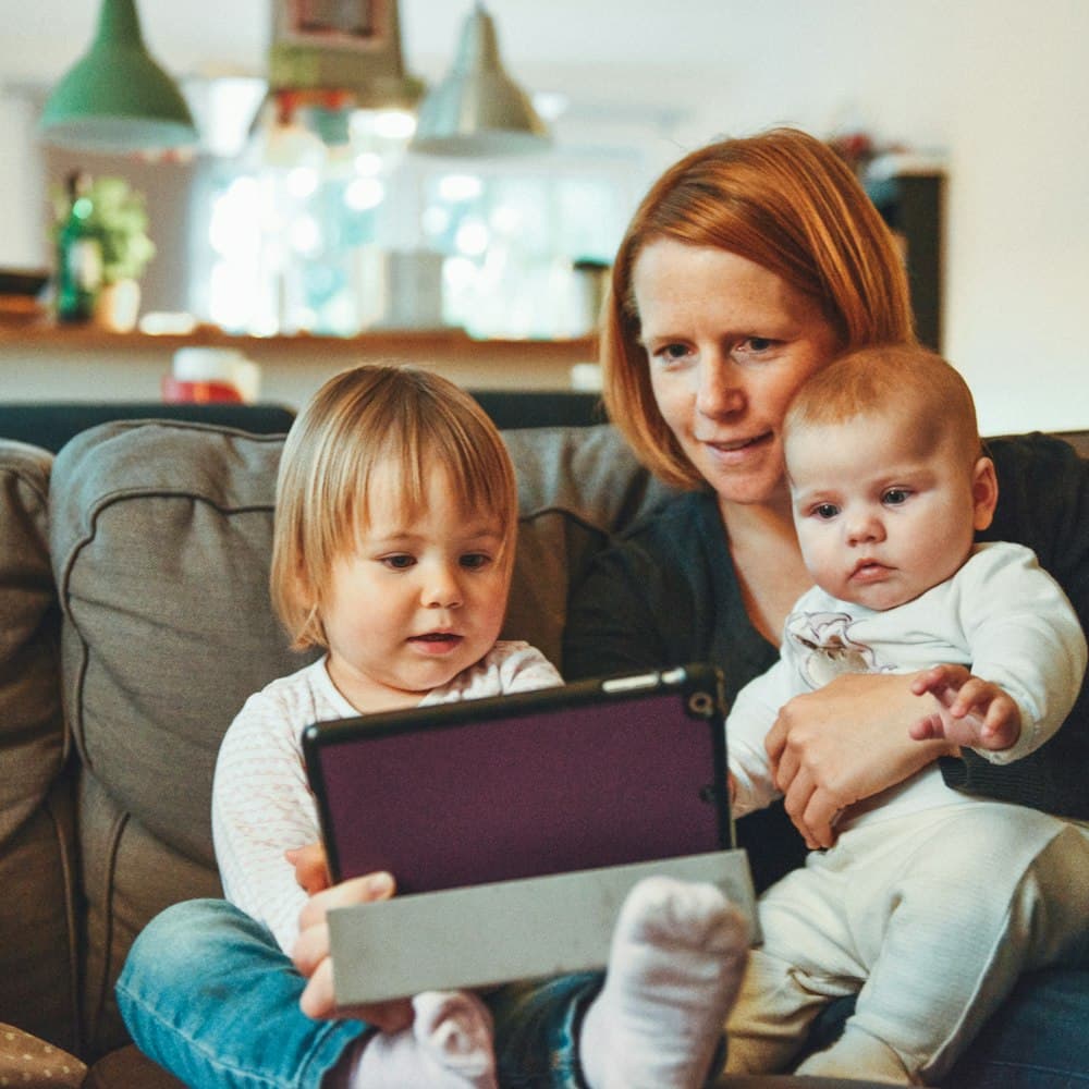 A mother on a sofa at home with her toddler and baby, looking at a tablet together